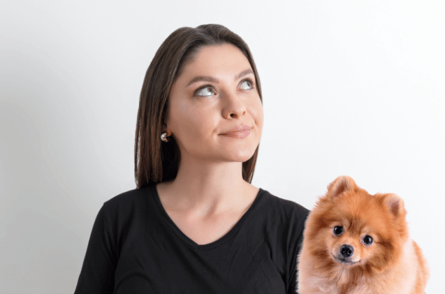 A woman in a black shirt gazes thoughtfully upward, seated next to her fluffy brown Pomeranian against a white background, perhaps pondering how to set up a pet business and how to get pet owner clients