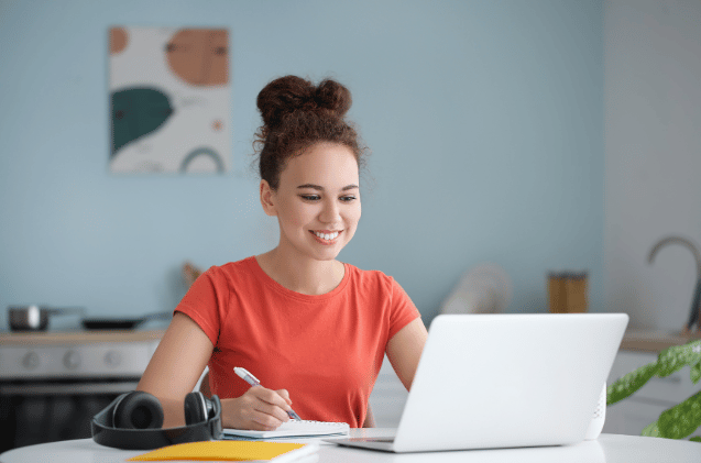 A woman in a red shirt is smiling at her laptop, researching how to set up a pet business. She jots down notes in her notebook, headphones beside her. The blurred kitchen and abstract wall art provide a cozy backdrop as she plans for her entrepreneurial journey. woman Improving Skills and Services at her dog walking and pet sitting business by doing training