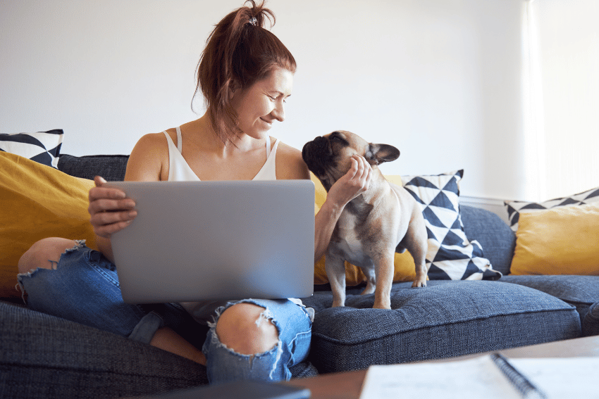 A woman with brown hair in a bun sits on a sofa, holding a laptop, while her French Bulldog rests its paw on her chest. The room exudes comfort with black and white patterned and yellow cushions. Nearby, a notebook and pen await. She's researching affordable pet boarding in Sydney for peace of mind.