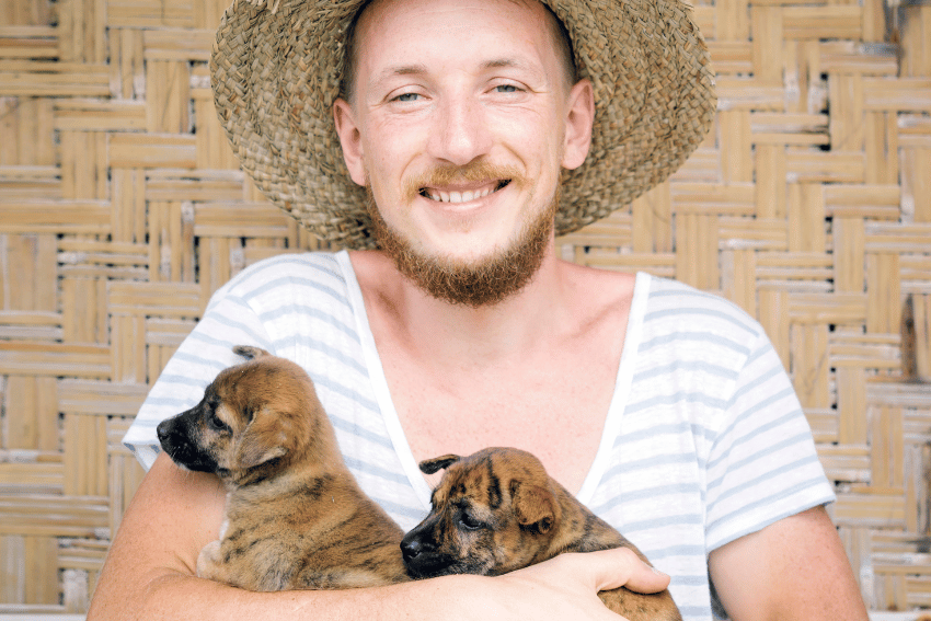 A person with a straw hat and striped shirt smiles while holding two small brown puppies, showing the joy of early socialization. They sit against a woven straw background, embodying the gentle art of introducing puppies to the world before vaccinations.