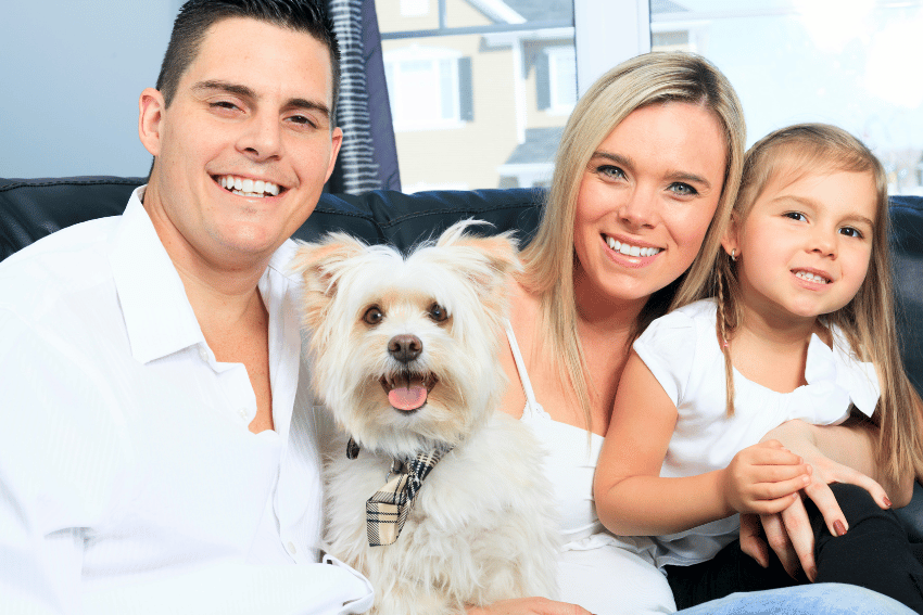 A smiling family sits on a couch with a fluffy white dog, wearing a plaid bowtie. The man, woman, and young girl in matching white tops bask in the cozy atmosphere. Behind them, a window reveals Melbourne's scenic homes, reminding them of the nearby affordable pet boarding options when they're away.